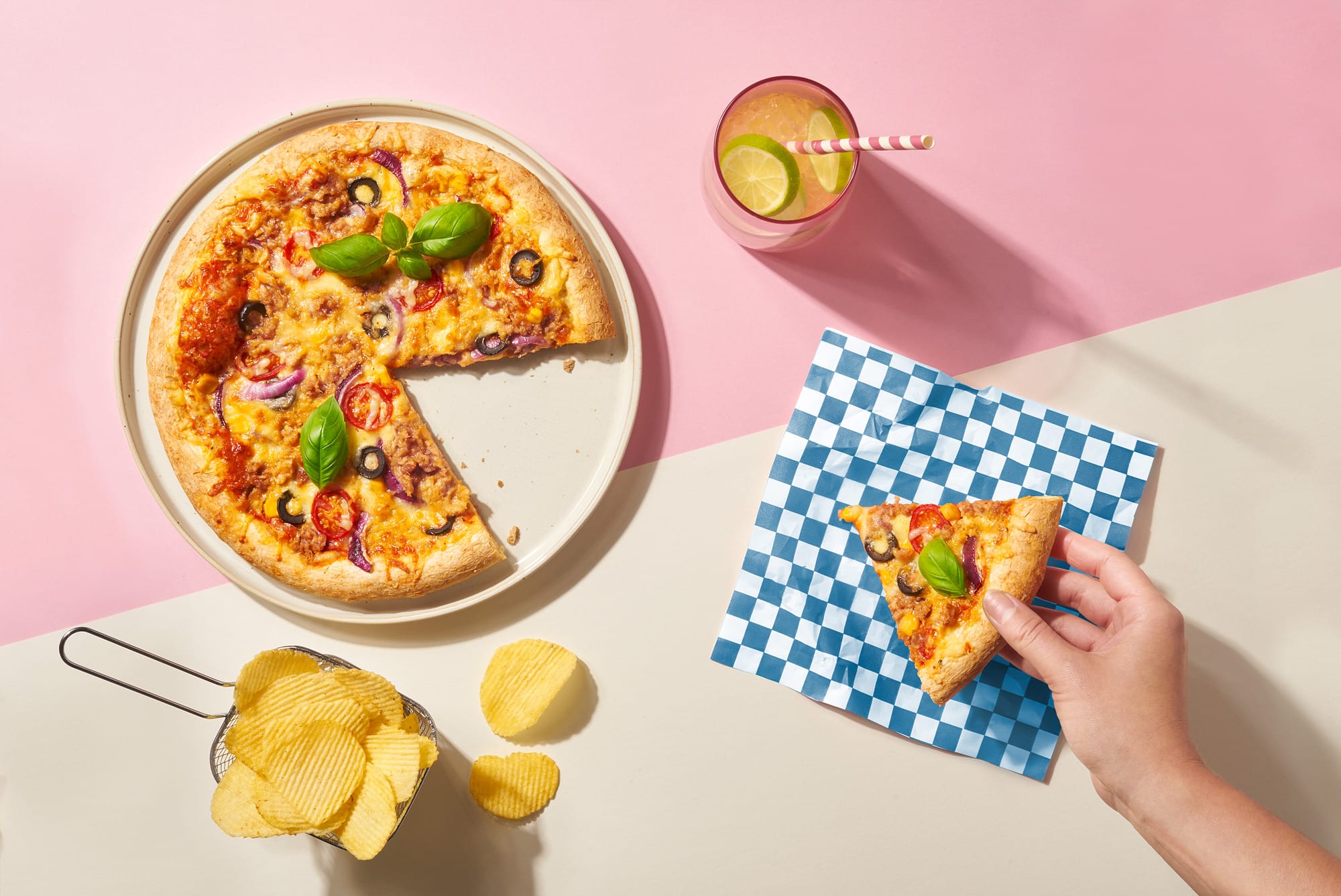 Lecker aussehende Pizza mit Thunfisch, Oliven, Tomaten und Zwiebeln, bei der ein Stück bereits gerade herausgenommen wird, daneben stehen Chips und ein Glas Limonade, Ansicht von oben