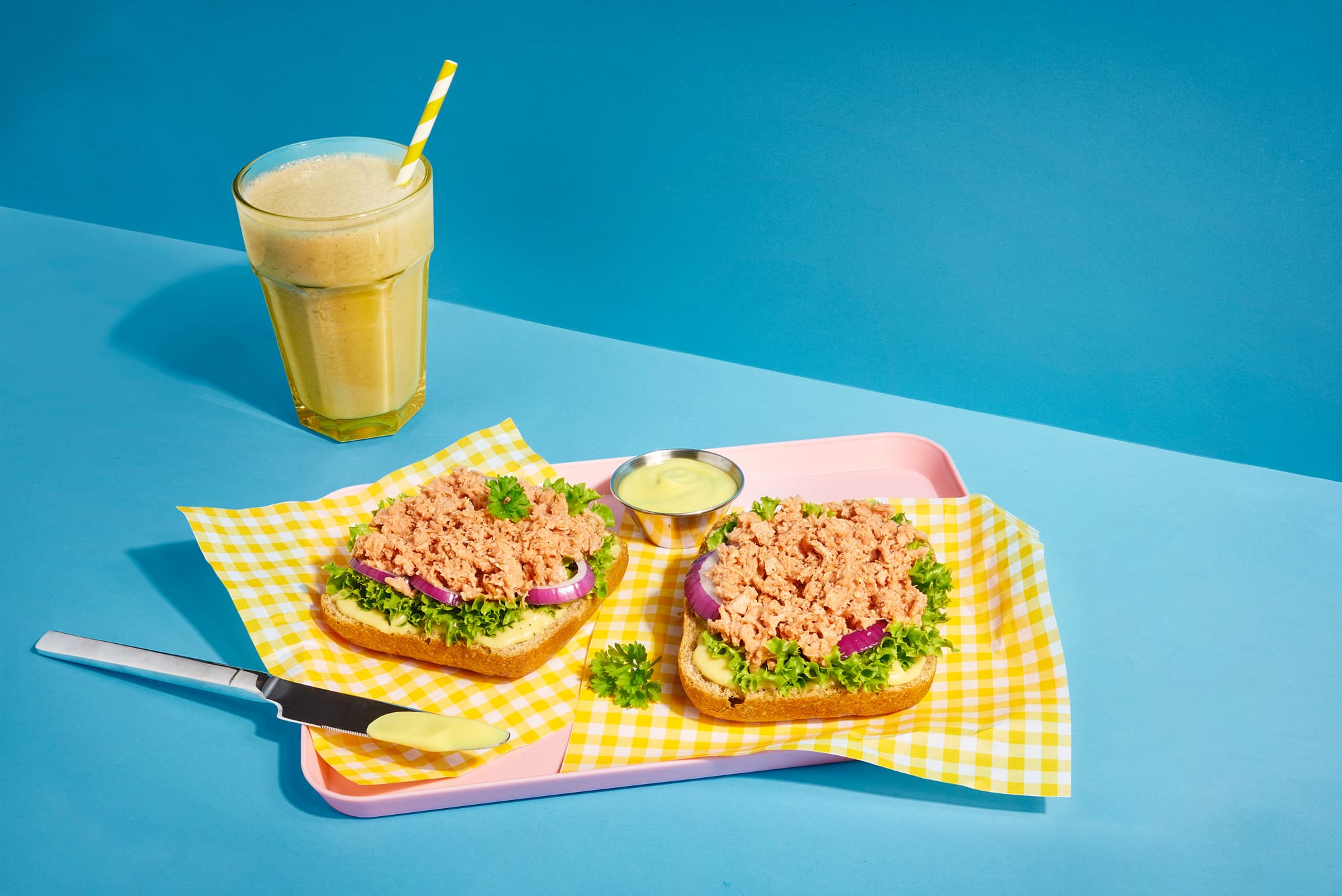 Zwei lecker angerichtete Thunfisch Toasts mit Zwiebeln und Salat, daneben steht ein gelber Smoothie